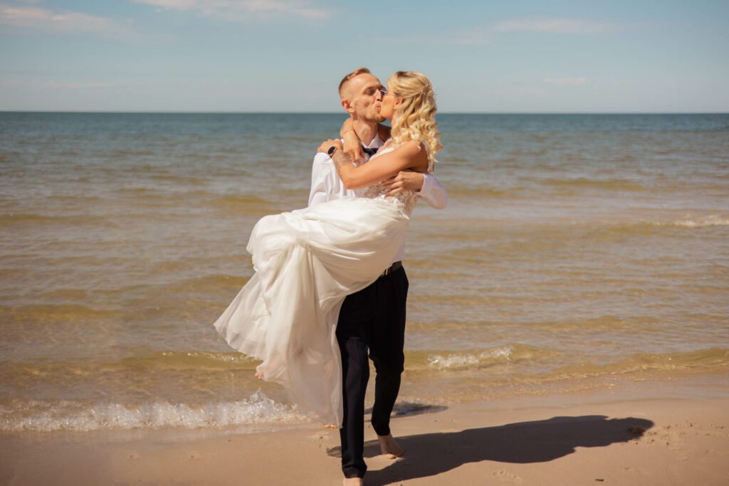 Pan młody niesie pannę młodą na rękach po plaży